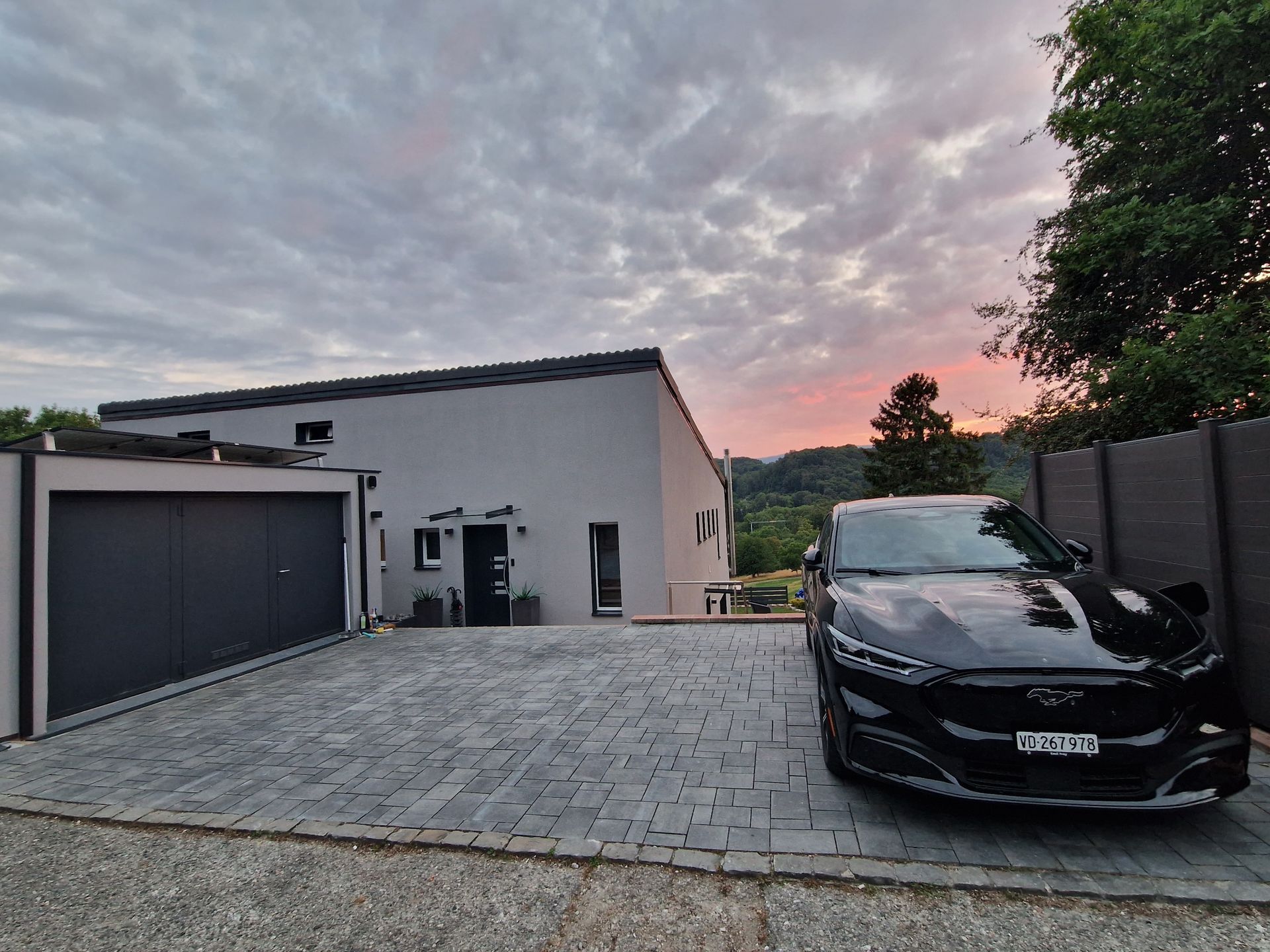 Maison grise moderne avec voiture noire garée devant, allée pavée de pierres grises. Ciel de coucher de soleil.