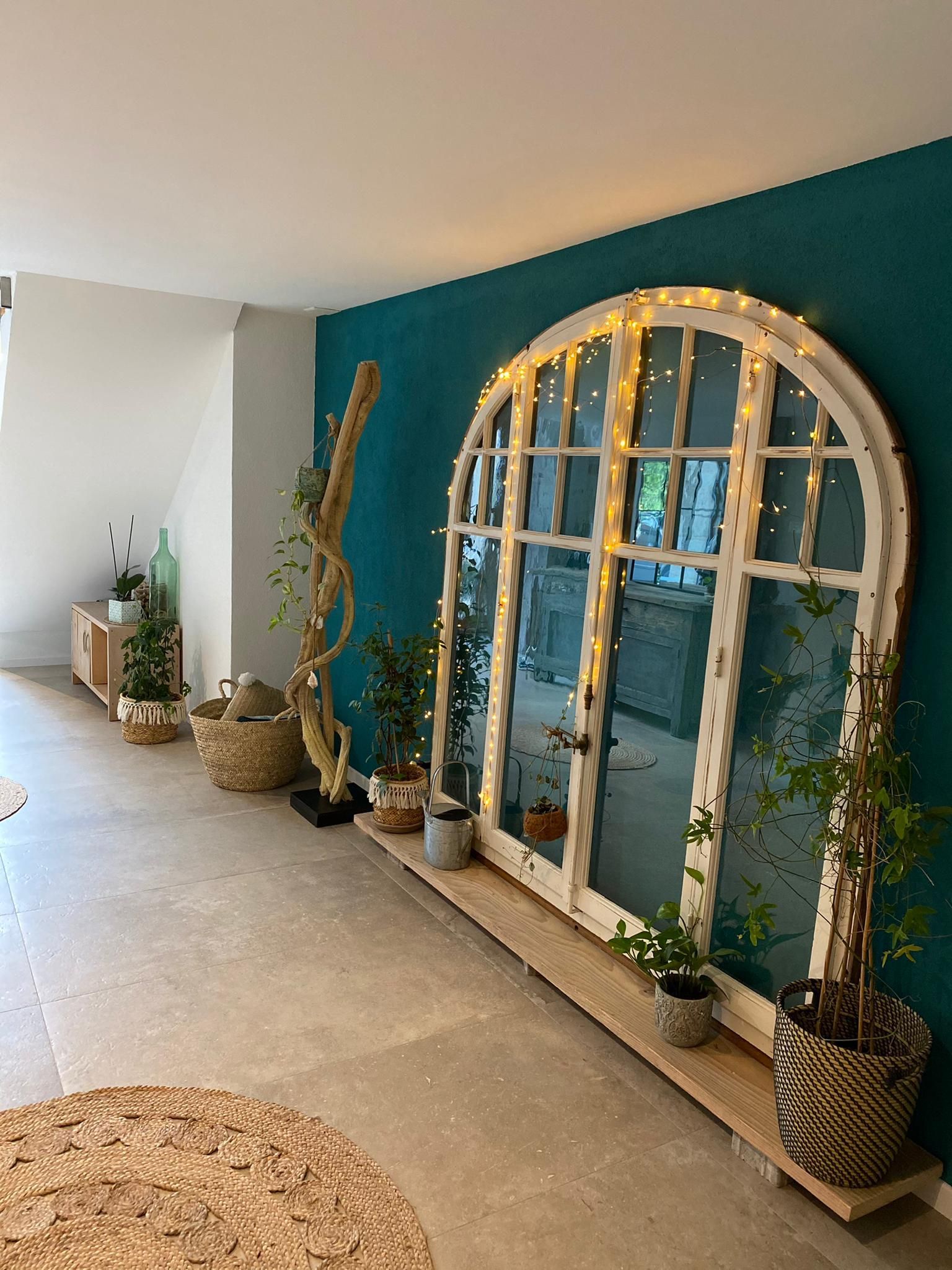 Grande fenêtre cintrée blanche ornée de guirlandes lumineuses sur un mur turquoise, des plantes et une estrade en bois clair.