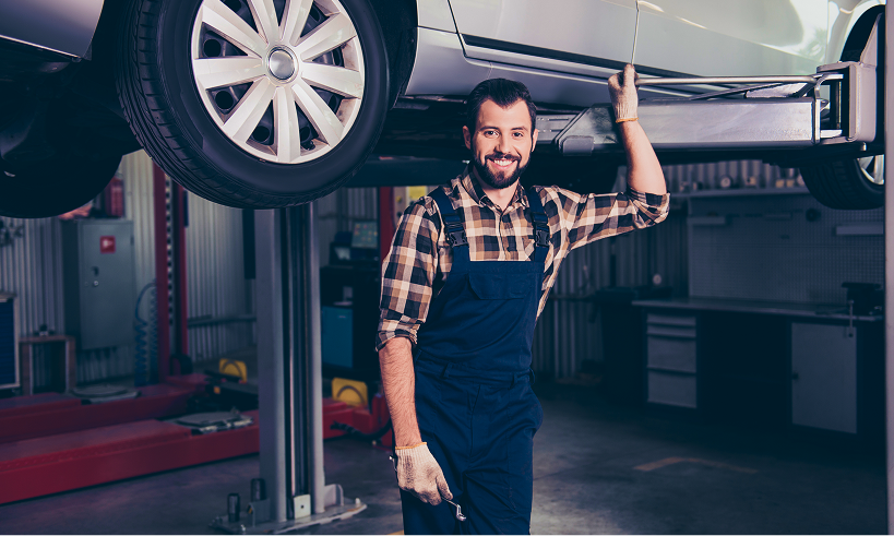 Mechanic in overalls smiles under a raised car in a garage, holding a wrench. | Doran-King Garage Inc