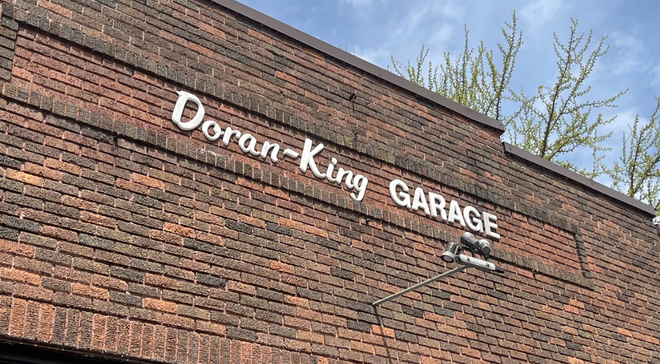 Mechanic in overalls smiles under a raised car in a garage, holding a wrench. | Doran-King Garage Inc