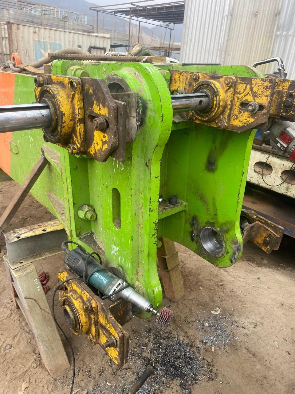 Construcción de metal verde que se trabaja al aire libre con una herramienta de esmerilado; detalles en amarillo/naranja.