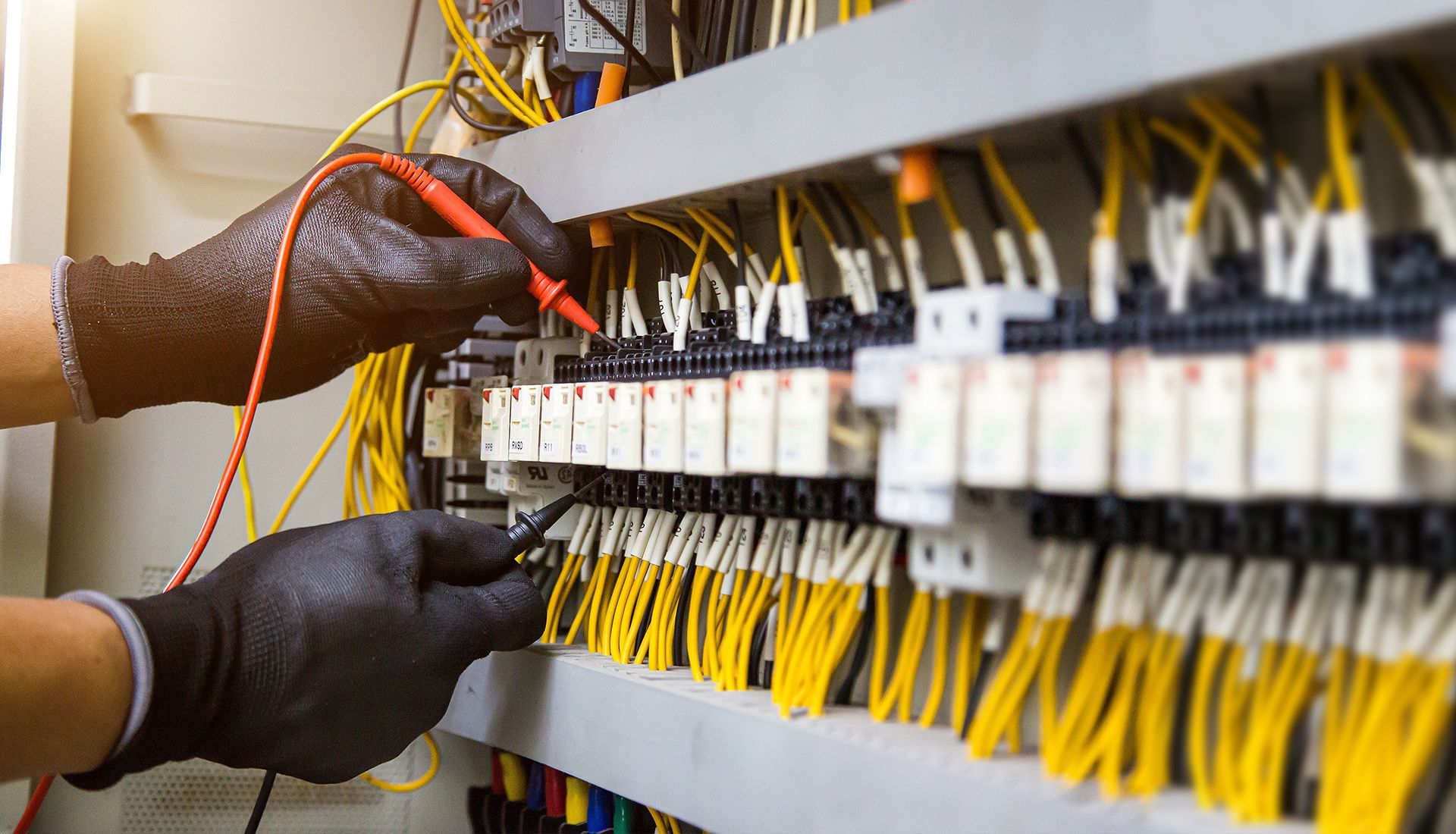 Hands with black gloves testing electrical wiring in a panel with a multimeter. Yellow wires prominent.