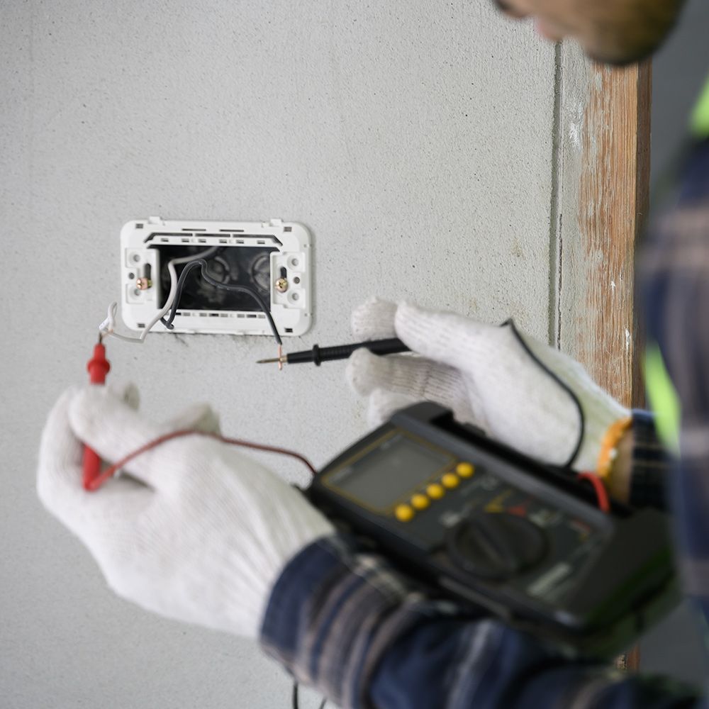 Electrician testing electrical outlet with a multimeter. White gloved hands, grey wall, black tool, red & black wires.