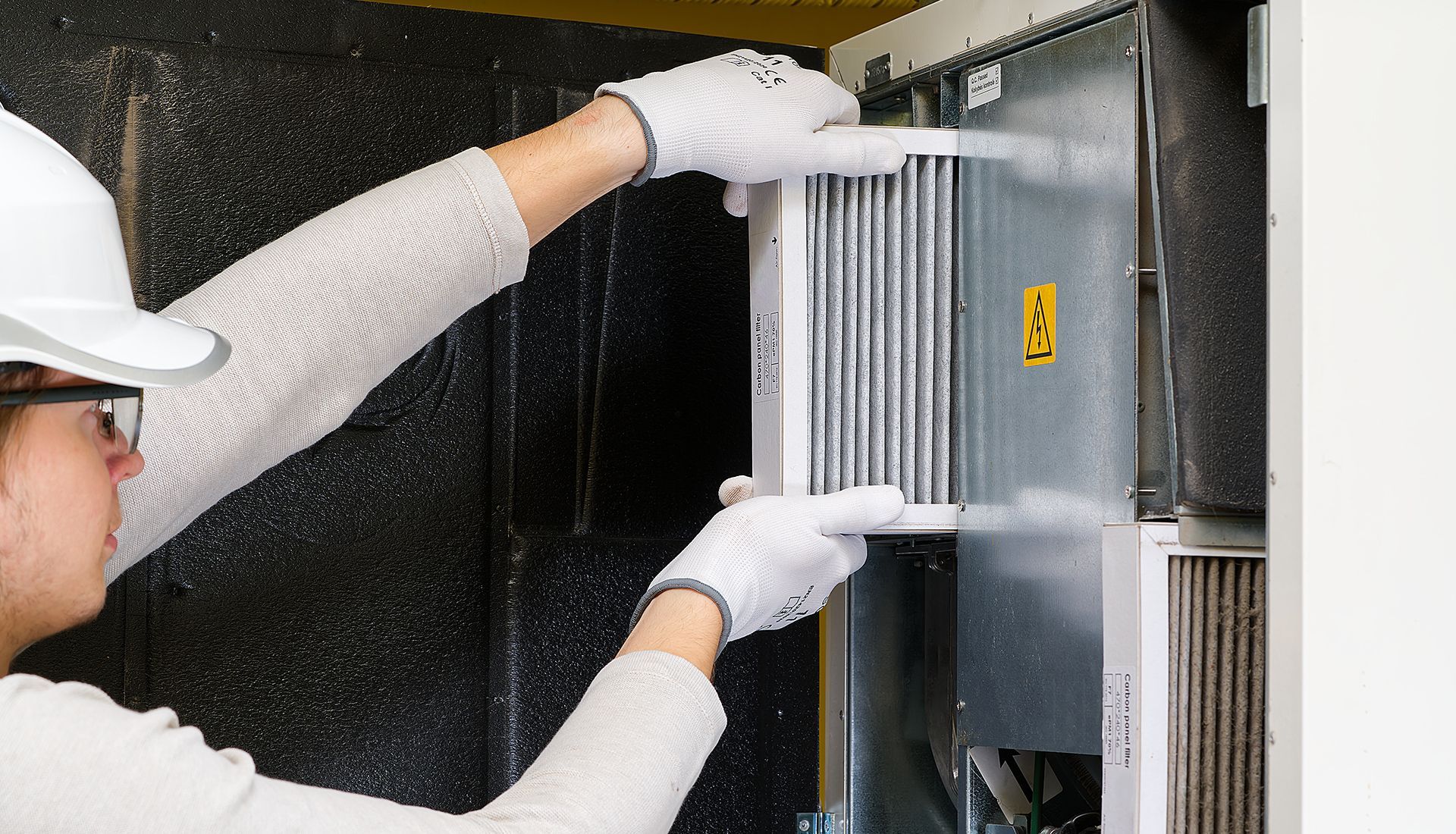 Person in a hard hat and gloves changing an air filter in a ventilation system.