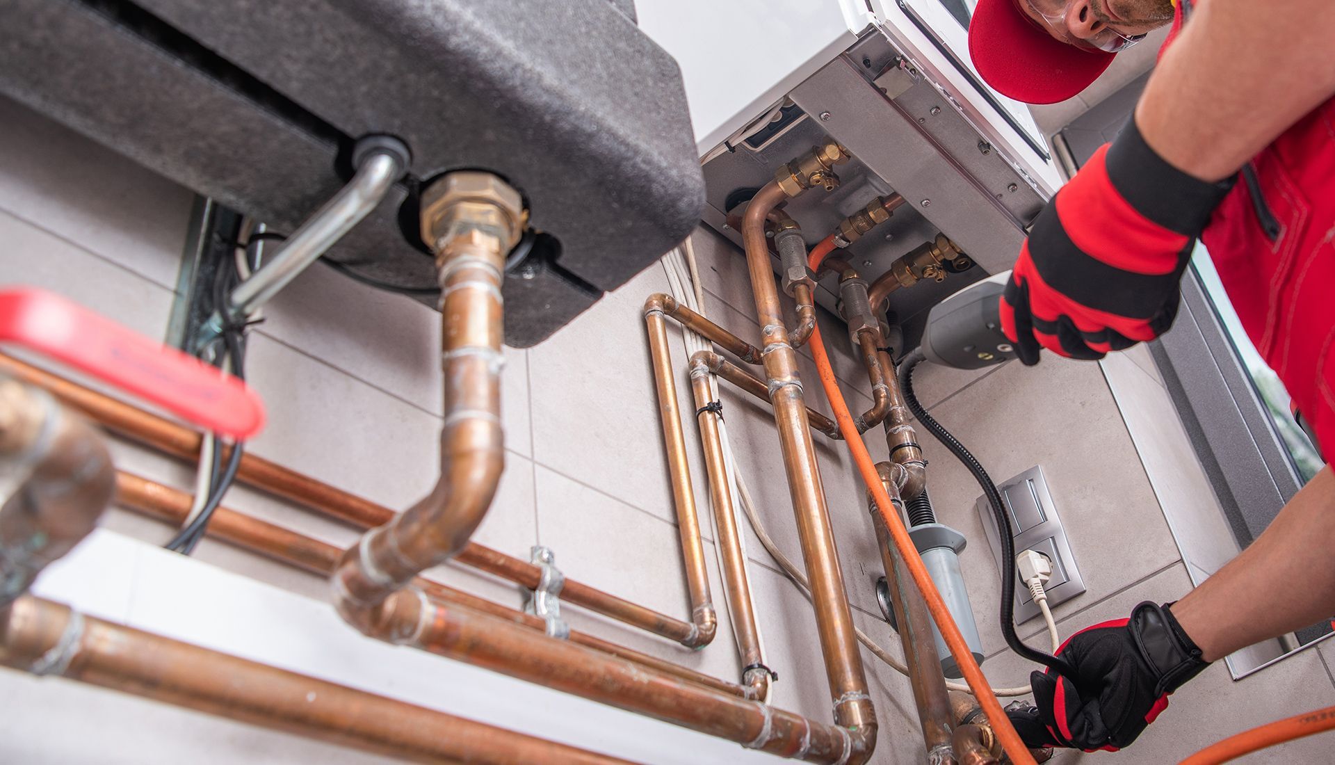 Plumber in red overalls repairs pipes and electrical connections near a wall-mounted boiler and tank.