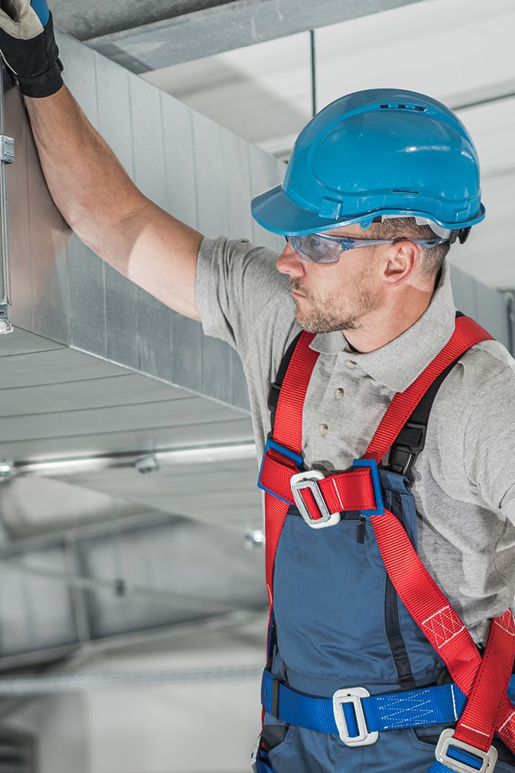 Construction worker in hard hat and safety harness, working on ductwork.