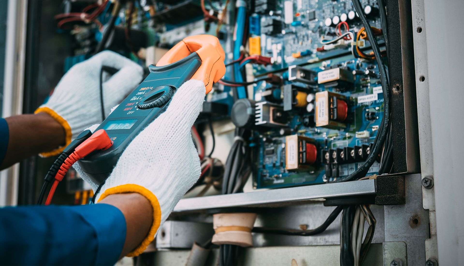 Person in gloves using a multimeter to inspect circuit board inside electrical equipment.