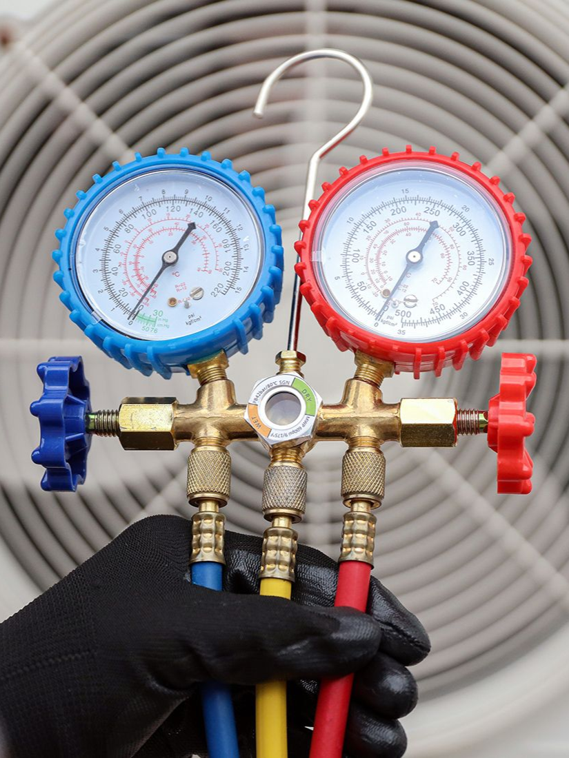 Hand holding HVAC manifold gauge set; gauges with blue and red dials; yellow, blue, and red hoses connected.