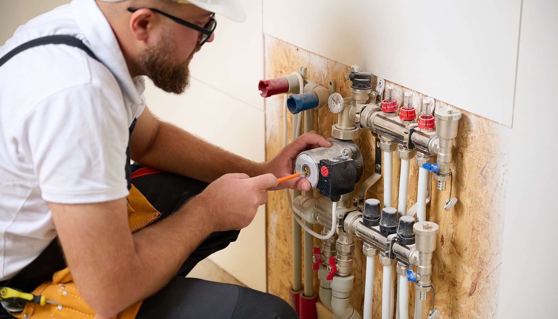 Plumber working on pipes, kneeling by a wall. He is using a screwdriver.