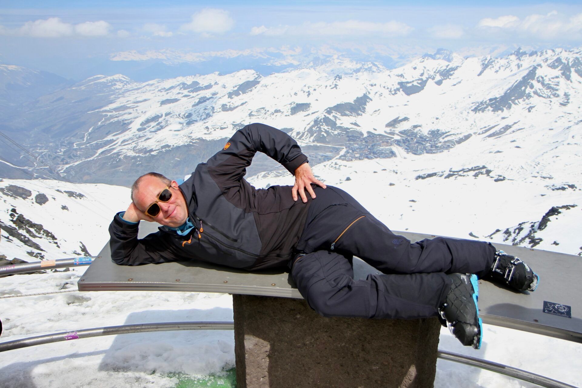 A man is laying on a bench on top of a snow covered mountain.