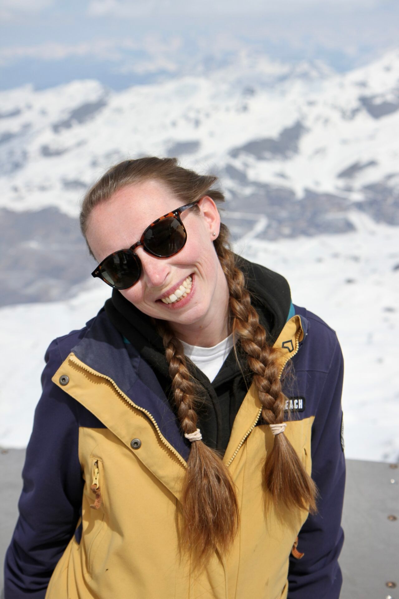 A woman wearing sunglasses and a blue jacket is smiling in front of a snowy mountain.