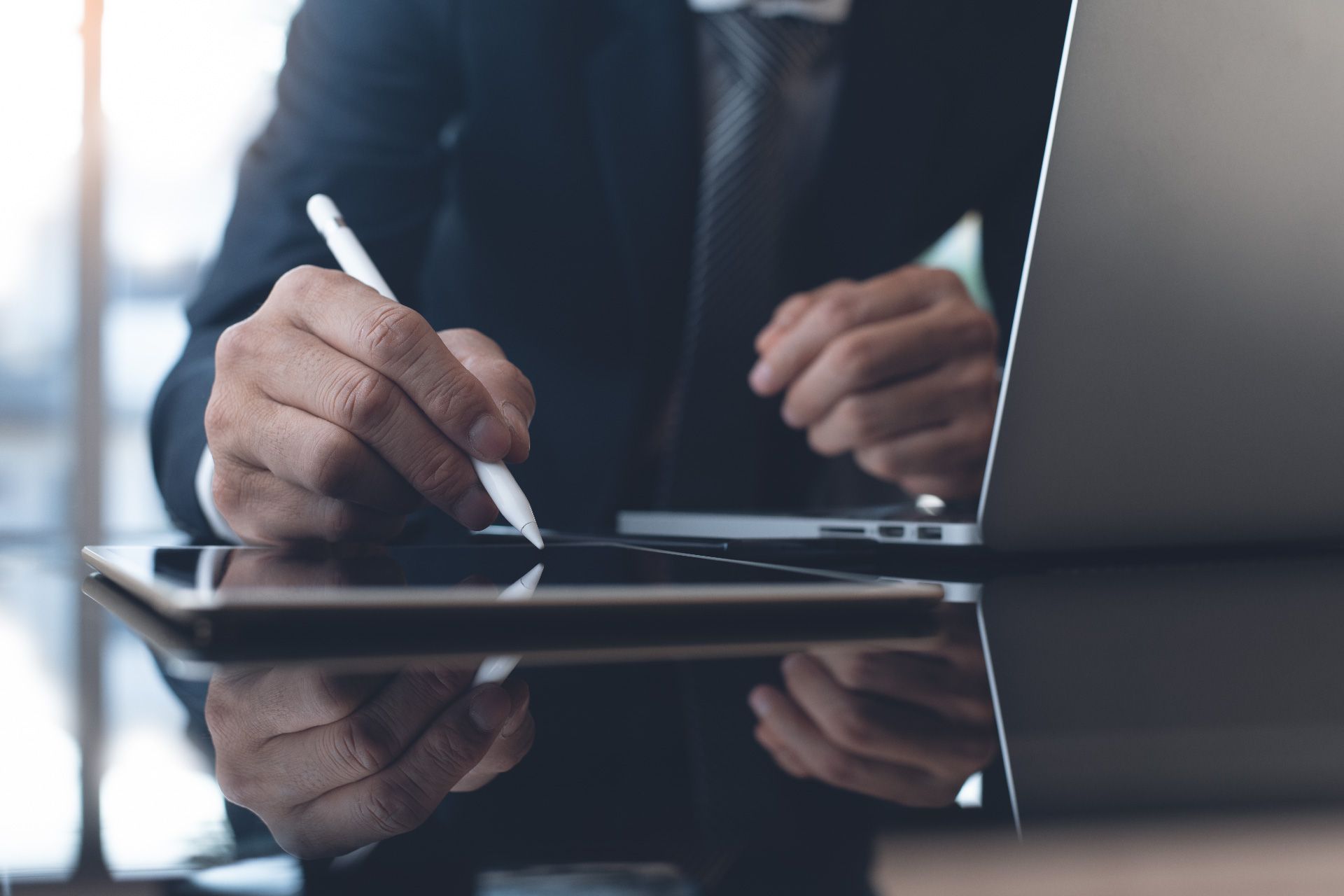 Hombre de traje firmando un documento en una tableta con un lápiz, con una computadora portátil al fondo, en una mesa reflectante.