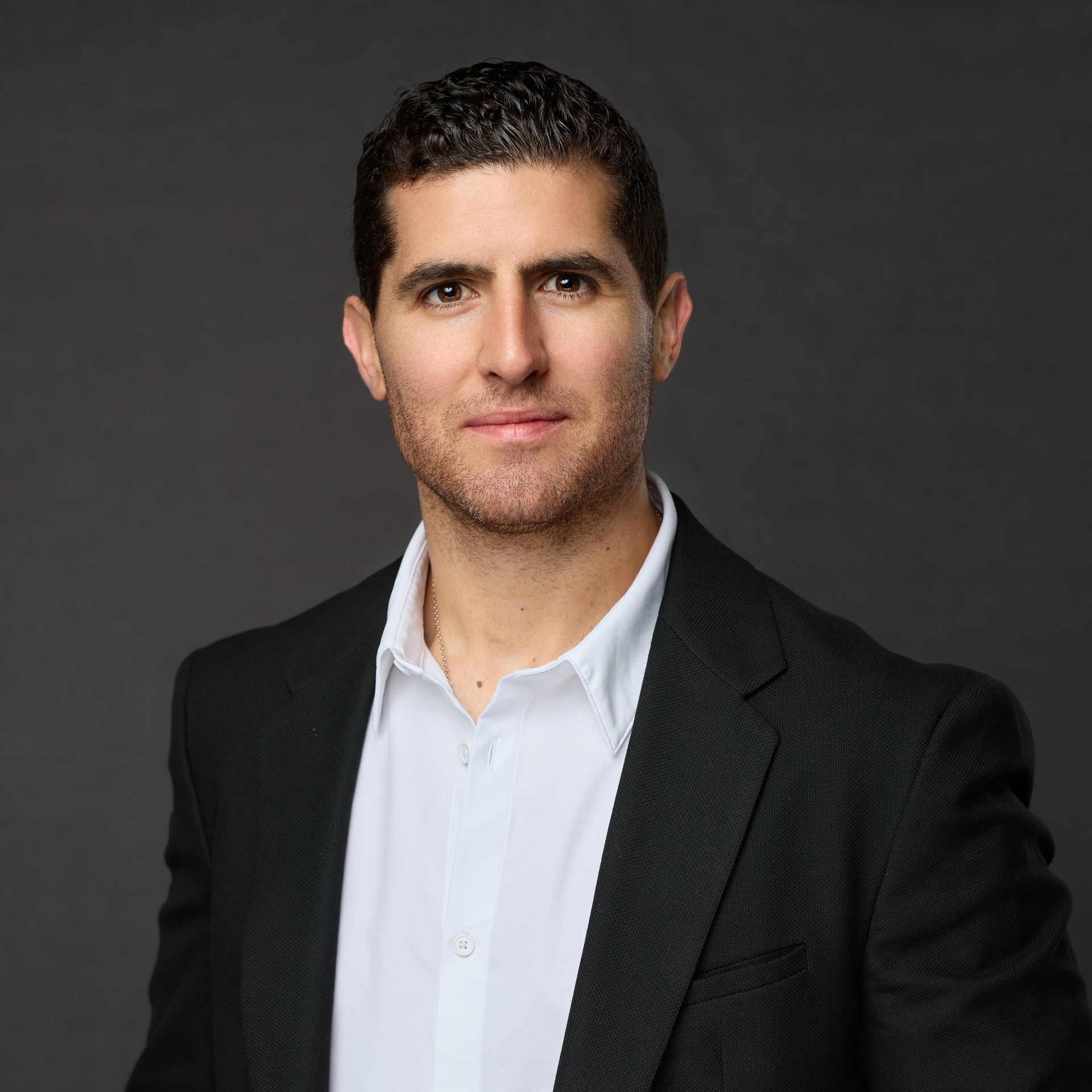 A man in a suit and white shirt is standing in front of a gray background.