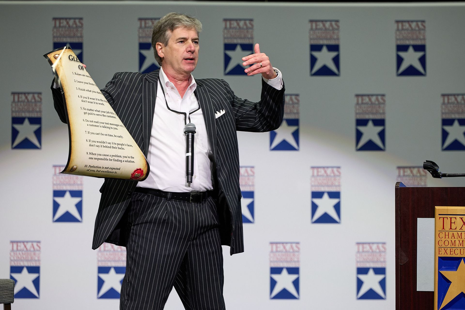 A man, who is the key note speaker at a conference is in a suit is holding a poster in front of a step and repeat. He 