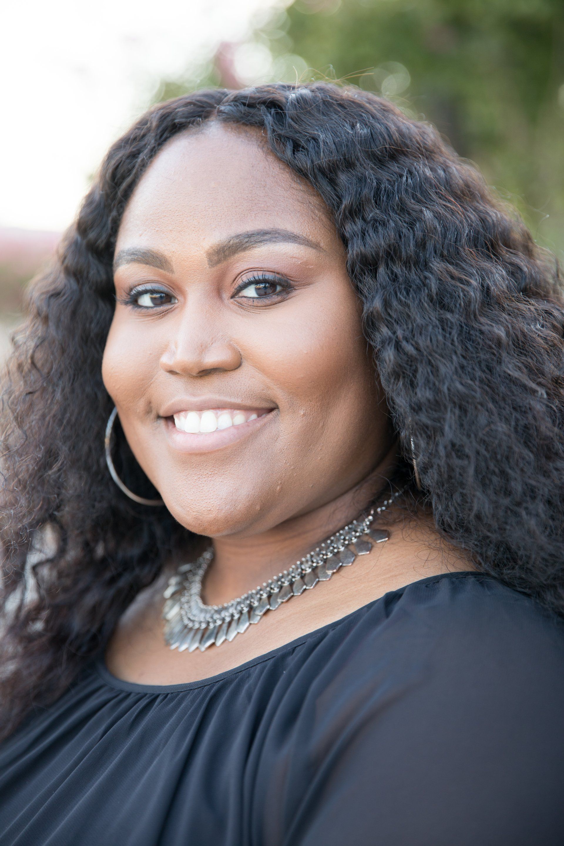 A woman with long curly hair wearing a black shirt and a necklace is smiling for the camera.