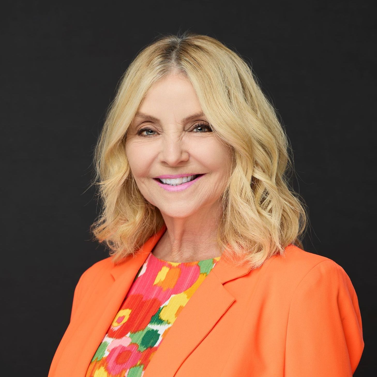 A woman wearing an orange jacket and a colorful shirt is smiling for the camera.