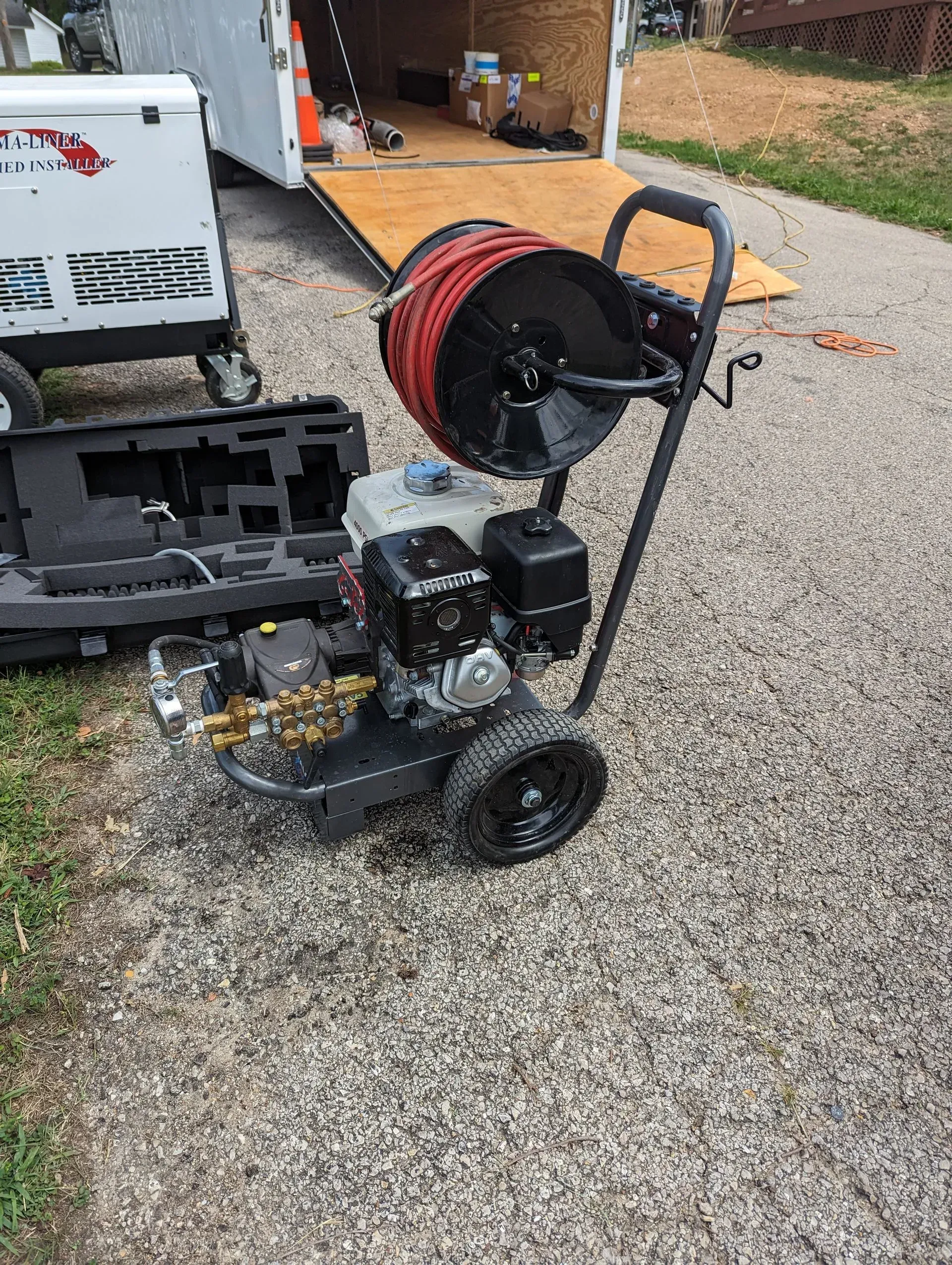 A pressure washer with a hose attached to it is sitting on a gravel driveway next to a trailer.