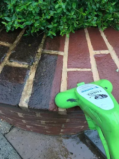 A bright green power washer nozzle cleans a section of dark, dirty brick, revealing clean, red brick underneath.