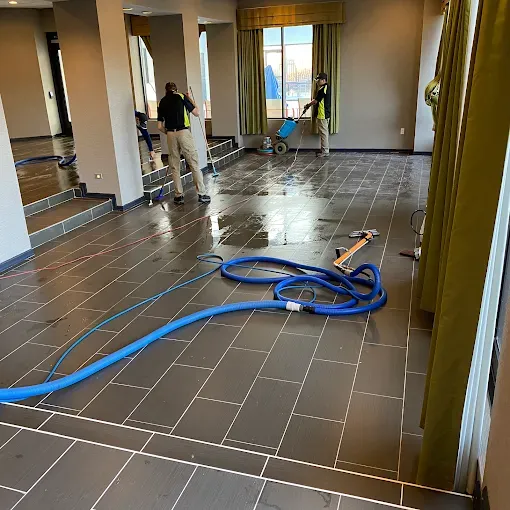 Two people in high-visibility vests clean a tiled floor with professional equipment in an indoor room.