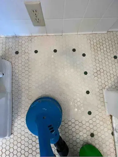 A person uses a blue floor scrubber to clean white hexagonal bathroom tiles with a dark accent border.