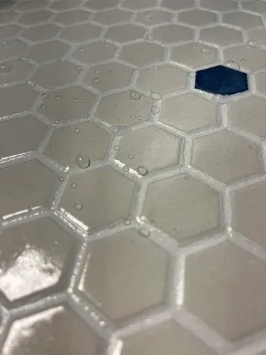 A close-up of light gray hexagonal floor tiles with a single dark blue accent tile, wet with scattered water droplets.