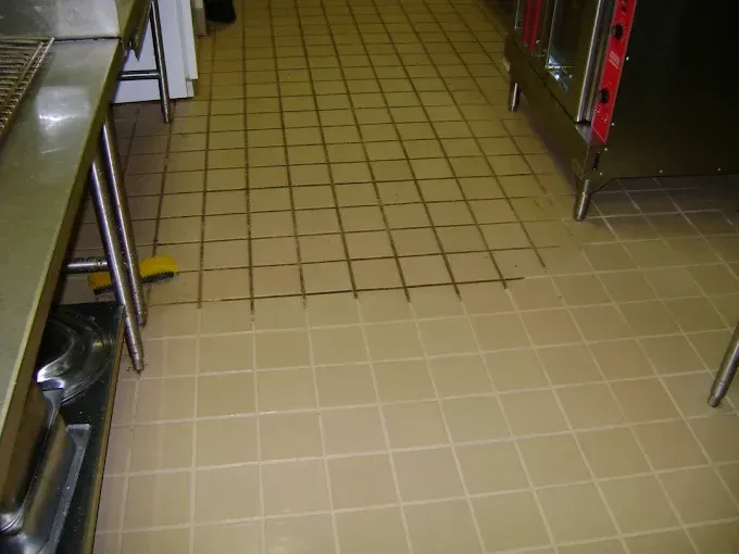 A commercial kitchen floor with a clean, light-colored tiled section meeting a darker, grime-filled tiled area.