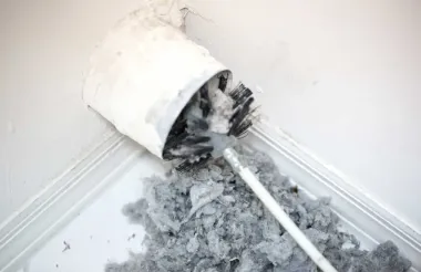 A dryer vent brush removing a large, dense clump of gray lint from a white wall corner.