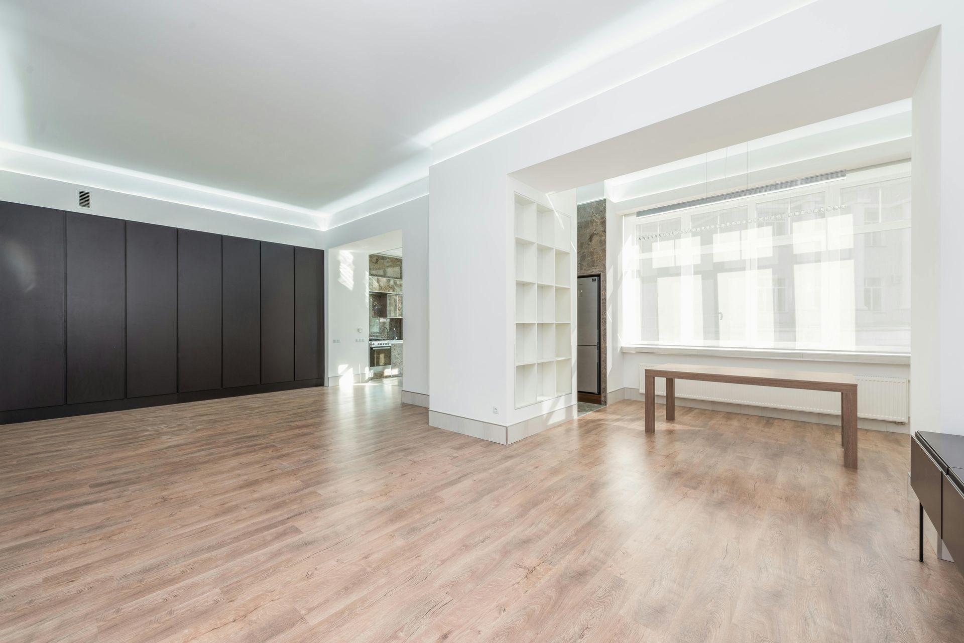 A bright, minimalist room featuring dark wood-paneled walls, light wood flooring, a white shelf unit, and a wooden table.