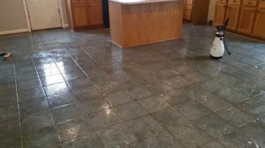 A brightly lit kitchen with gray stone-look floor tiles and a light wood kitchen island, with a spray bottle on the floor.