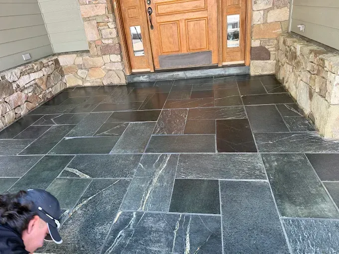 A person works on a newly installed dark stone patio in front of a house with a wood door and stone siding.