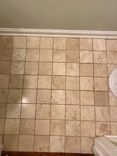 A tiled floor with beige, square natural stone tiles arranged in a grid pattern next to a white baseboard and toilet base.