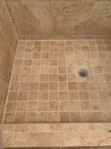 Shower floor tiled with small, tan stone-look squares, featuring a central circular drain and a tiled threshold.
