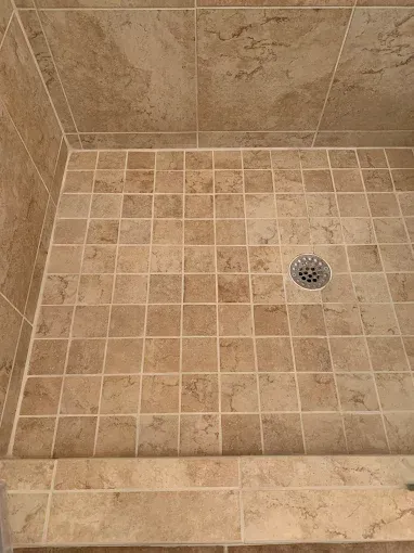 Shower floor tiled with small, tan stone-look squares, featuring a central circular drain and a tiled threshold.
