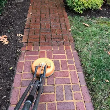 A surface cleaner pressure washes a red brick walkway between a mulch bed and a grassy lawn.