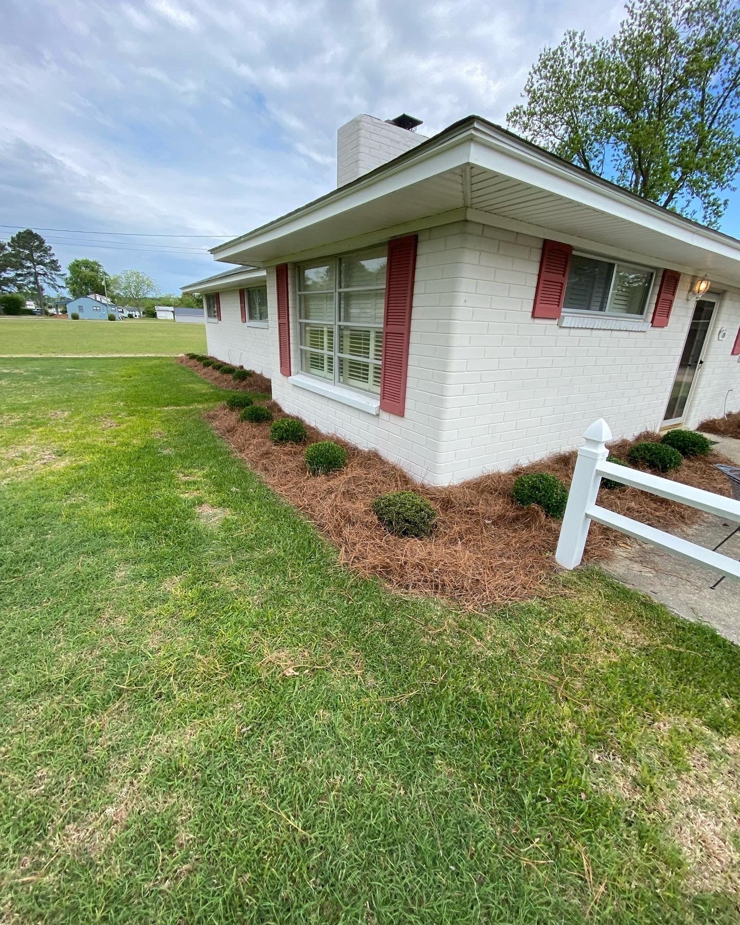 A white brick house with red shutters and a white fence in front of it.