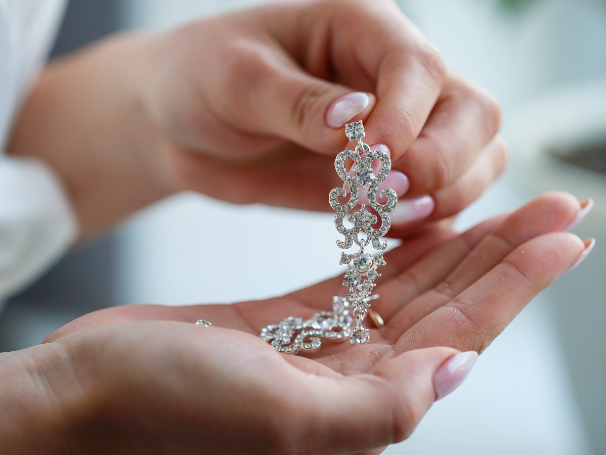 Hands holding an ornate, jeweled earring. The earring is silver, likely a bridal accessory.
