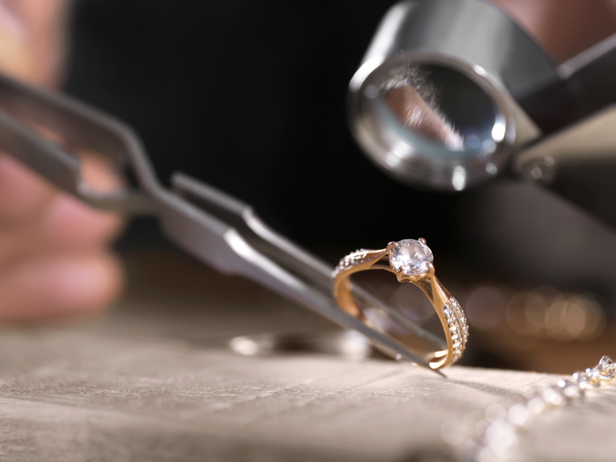 Jeweler inspecting a gold diamond ring with tweezers and magnifying glass.
