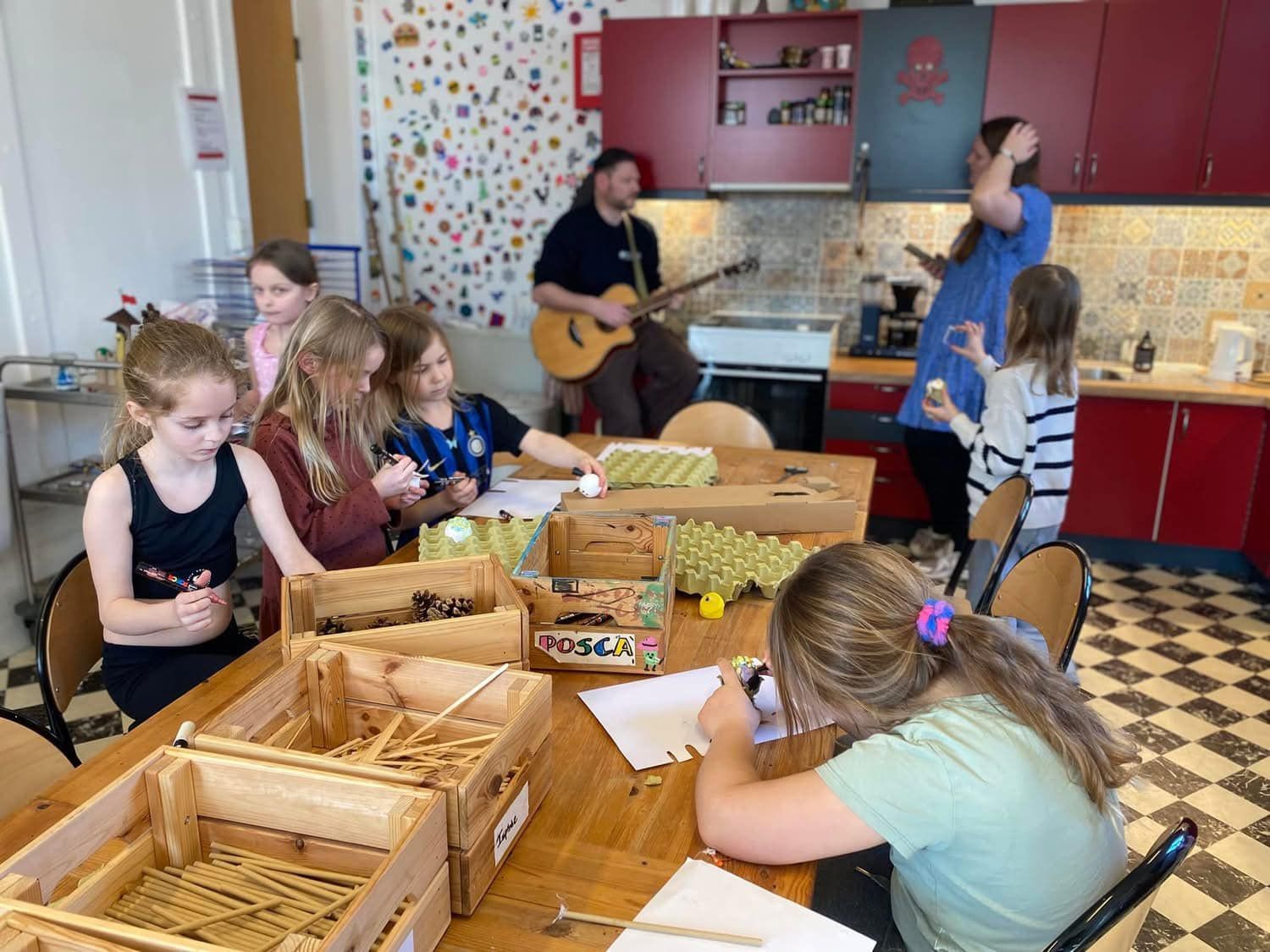 En gruppe børn sidder ved et bord i et køkken med en mand, der spiller guitar.