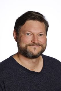 En mand med skæg har en blå sweater på og smiler til kameraet.