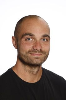 En mand med skæg har en sort skjorte på og smiler til kameraet.