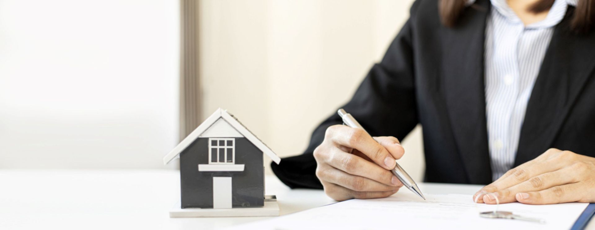 Person in business attire signing paperwork beside a model house, representing property documents.