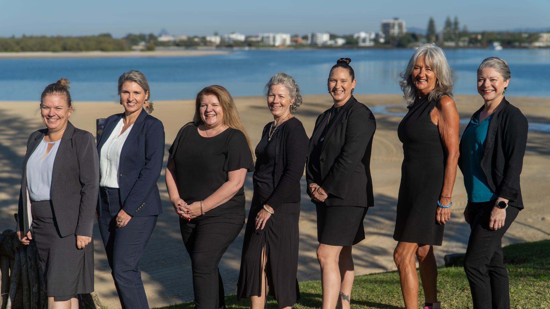 Seven women in business attire pose by a beach, smiling.