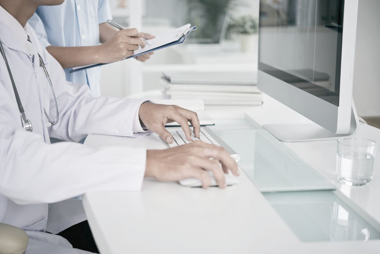 A doctor is typing on a computer keyboard while an assistant looks at a clipboard.