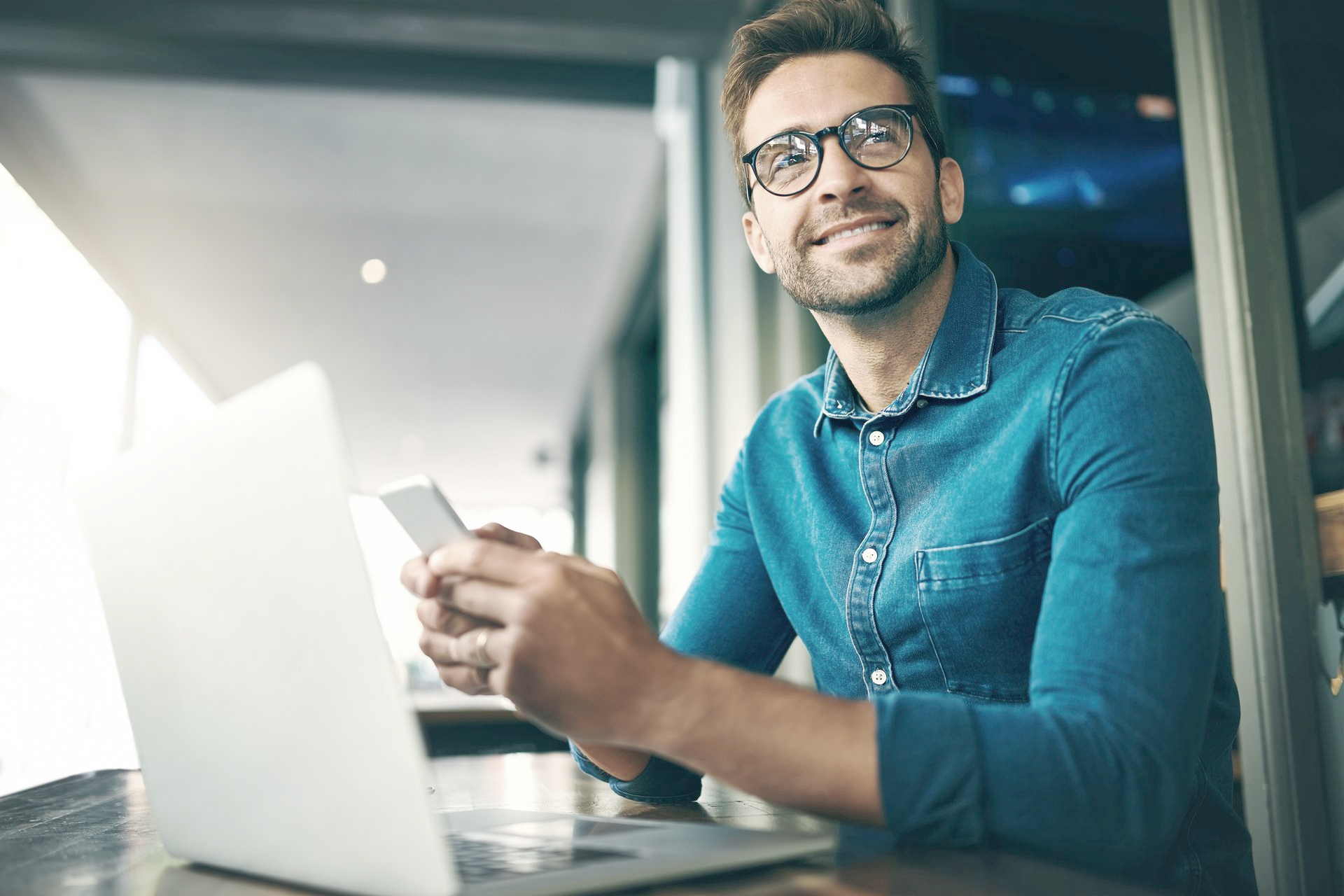 Man in blue shirt smiles while holding a phone and looking up, with a laptop open in front of him.