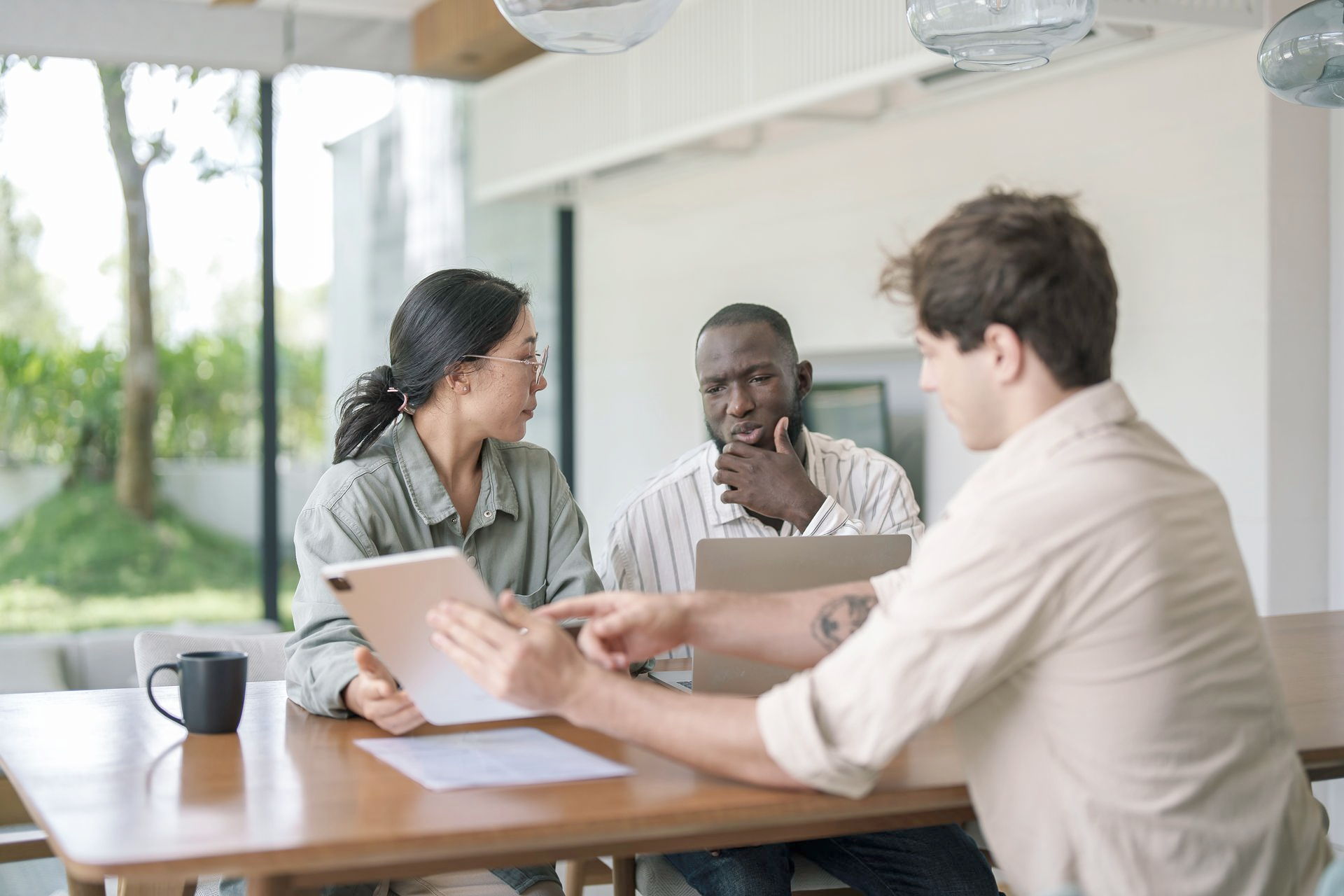 Three people at a table, looking at a tablet and laptop, discussing, inside a well-lit room.