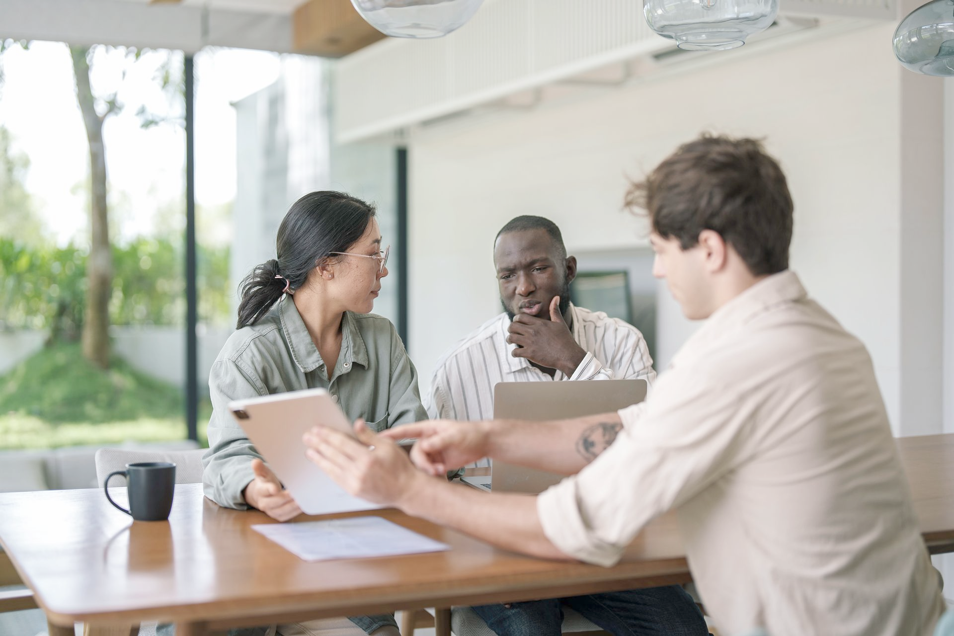Three people at a table, looking at a tablet and laptop, discussing. A man points at the screen.