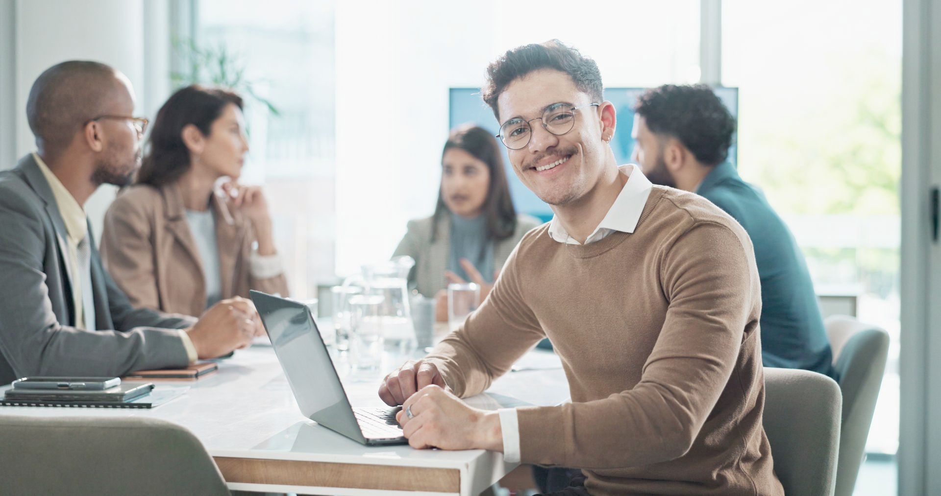 A man wearing glasses is smiling and sitting at a table with a laptop in front of a group of people.