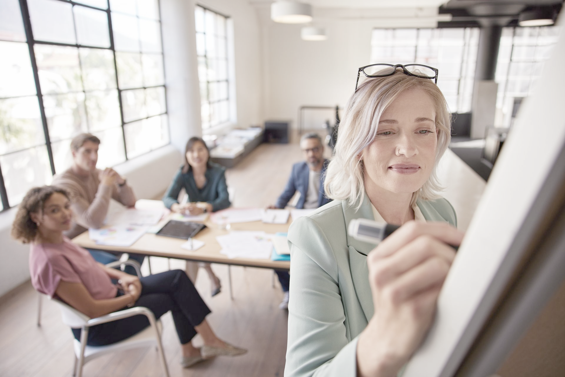 Woman in office drawing on whiteboard; team in background.