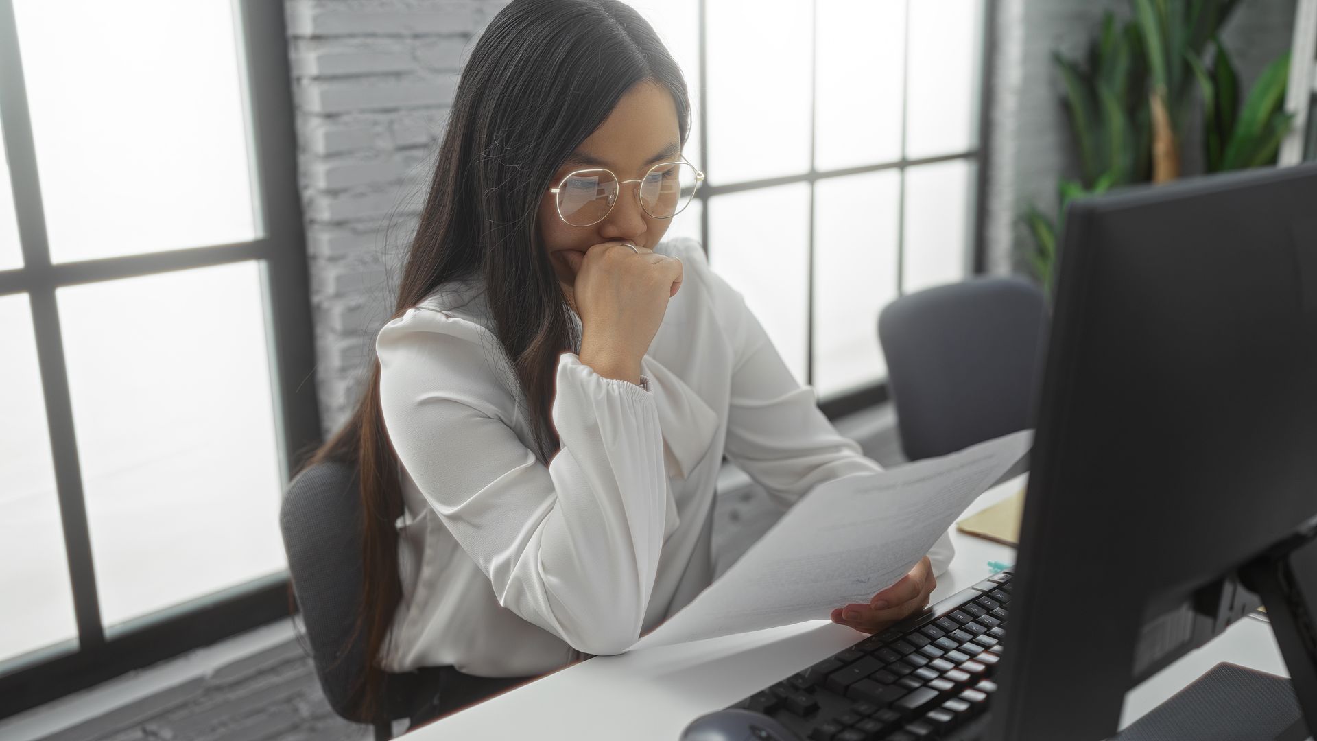 A person in a white top sits at a desk, looking at a document and a computer screen in a bright, modern office.