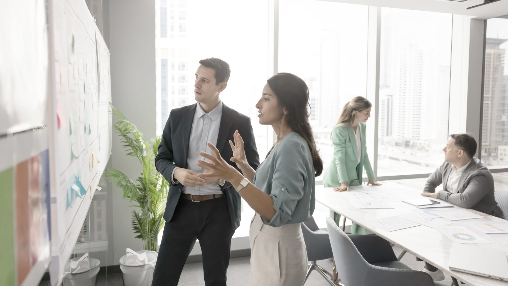 People in office discussing data on whiteboard. Sunlight streams in from a window.